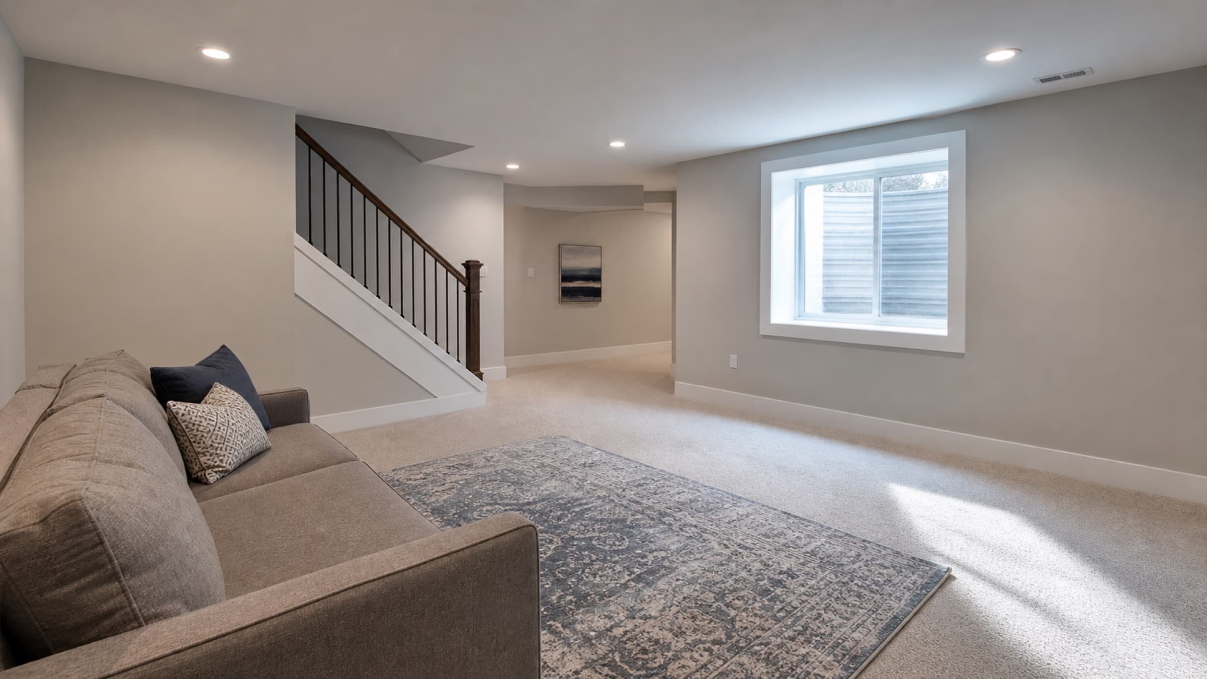 A finished basement family room with recessed lighting and a freshly painted stair stringer, staged sparsely