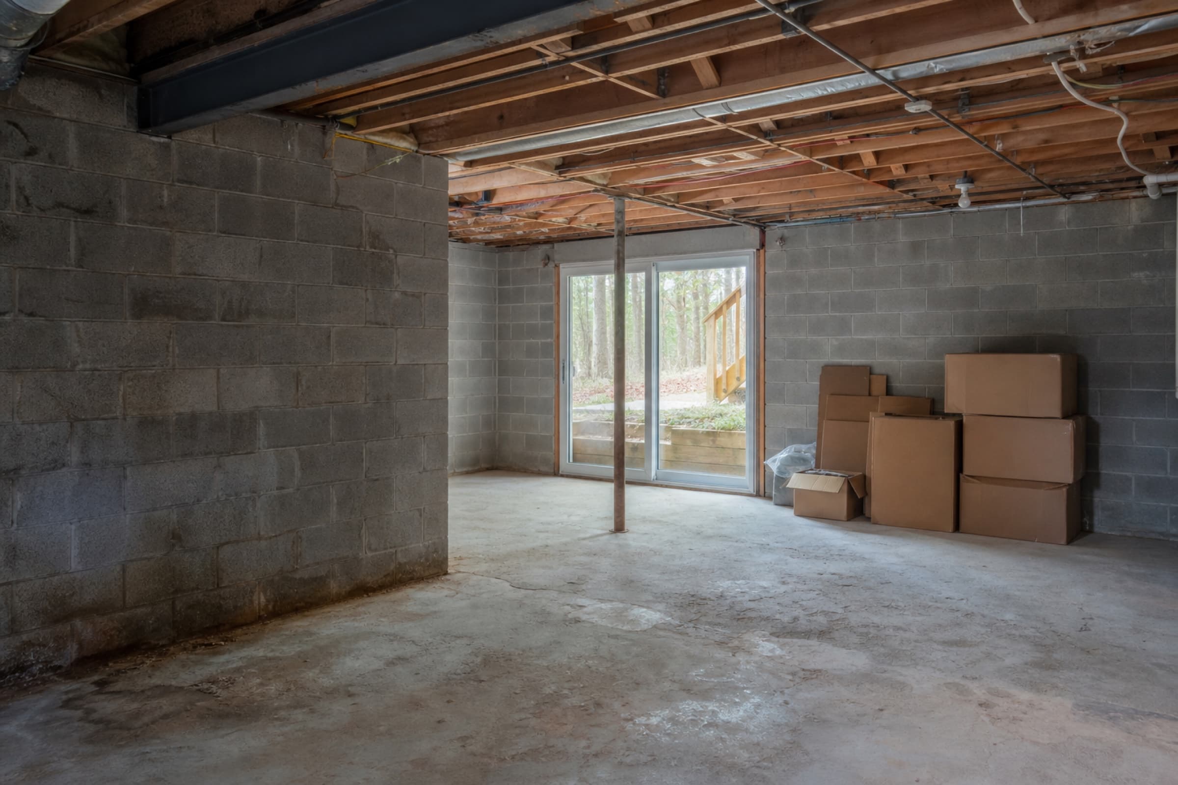 An unfinished basement with exposed joists, bare concrete and stored boxes along one wall