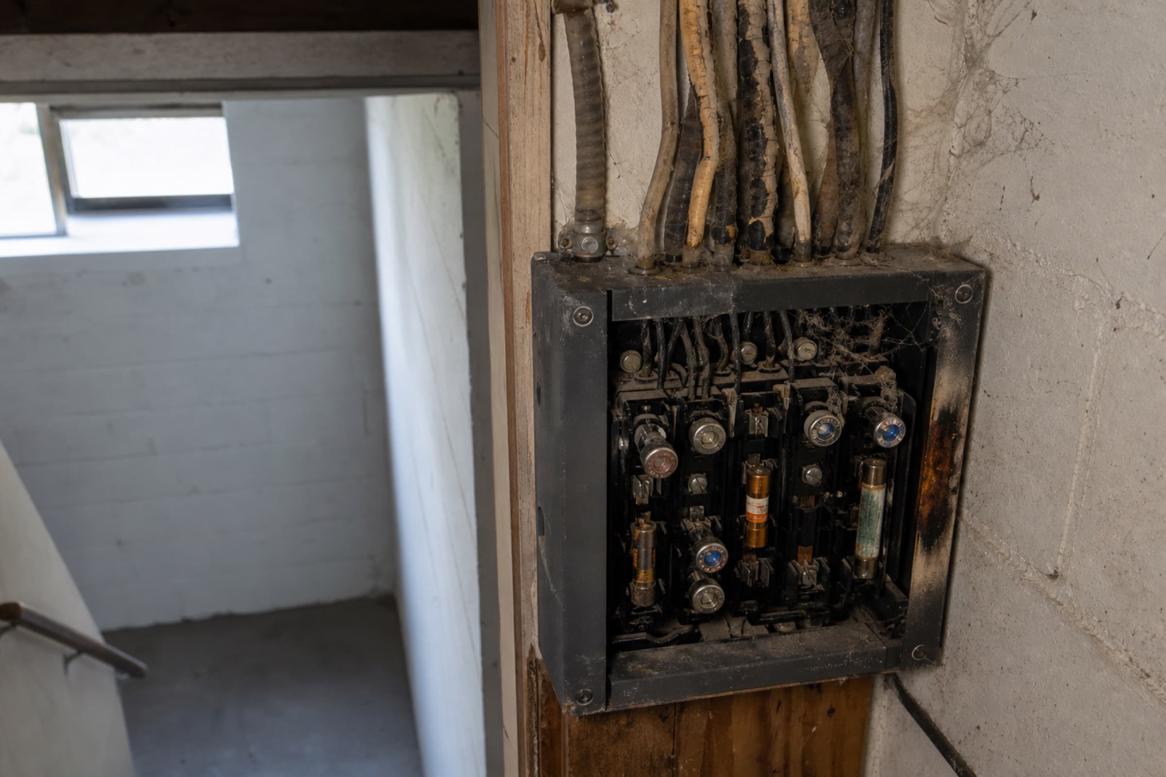 An original fuse panel with cloth-sheathed conductors and mismatched fuses in a basement stairwell