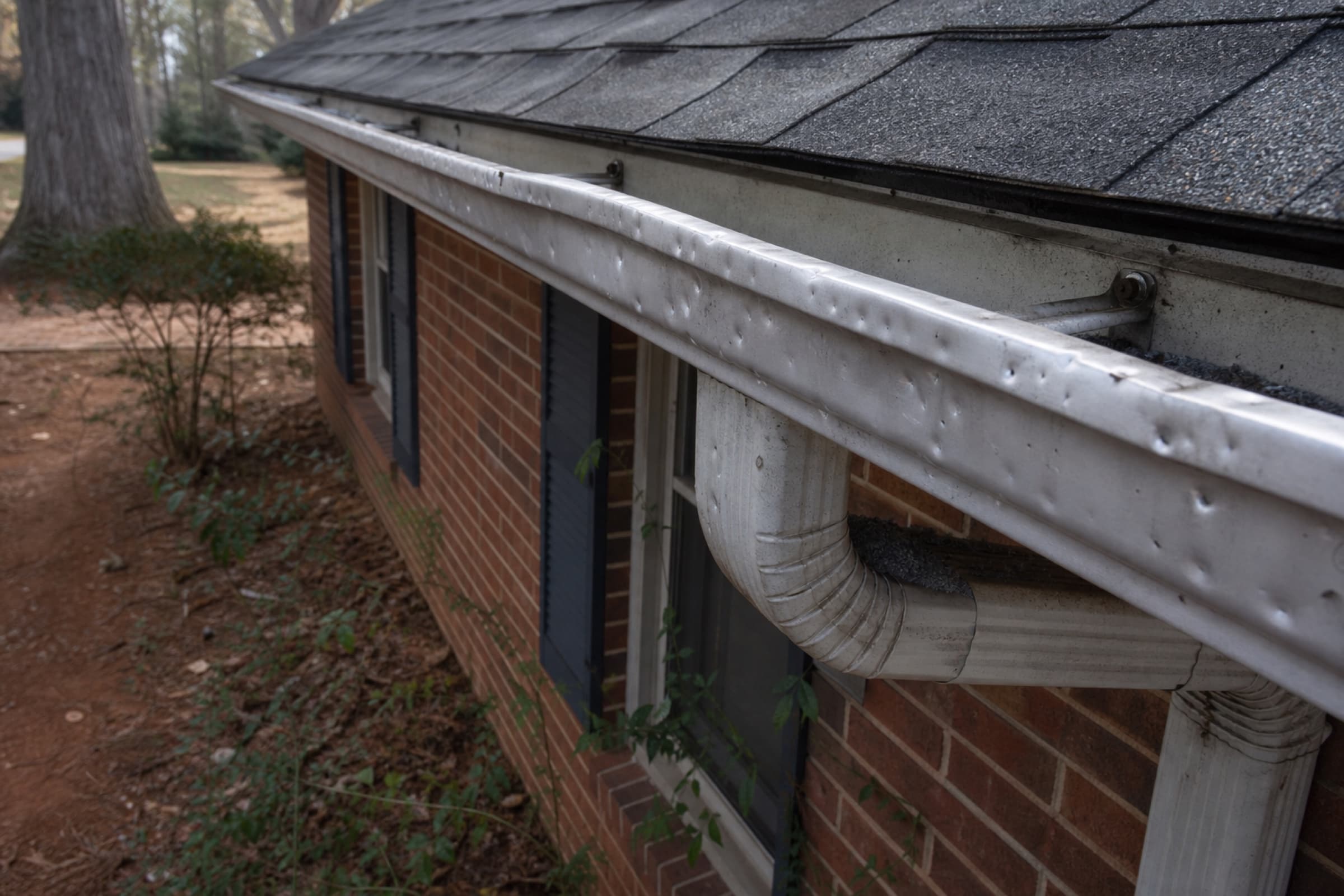 A dented aluminium gutter with shingle granules collected in the splash pan below the downspout
